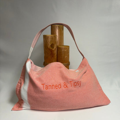 A dusty pink canvas shoulder bag with white side panels and a pink-and-white striped strap, featuring the text 'Tanned & Tipsy' embroidered in orange on the front. The bag is styled with three rustic wooden cylinder props against a draped white fabric backdrop.