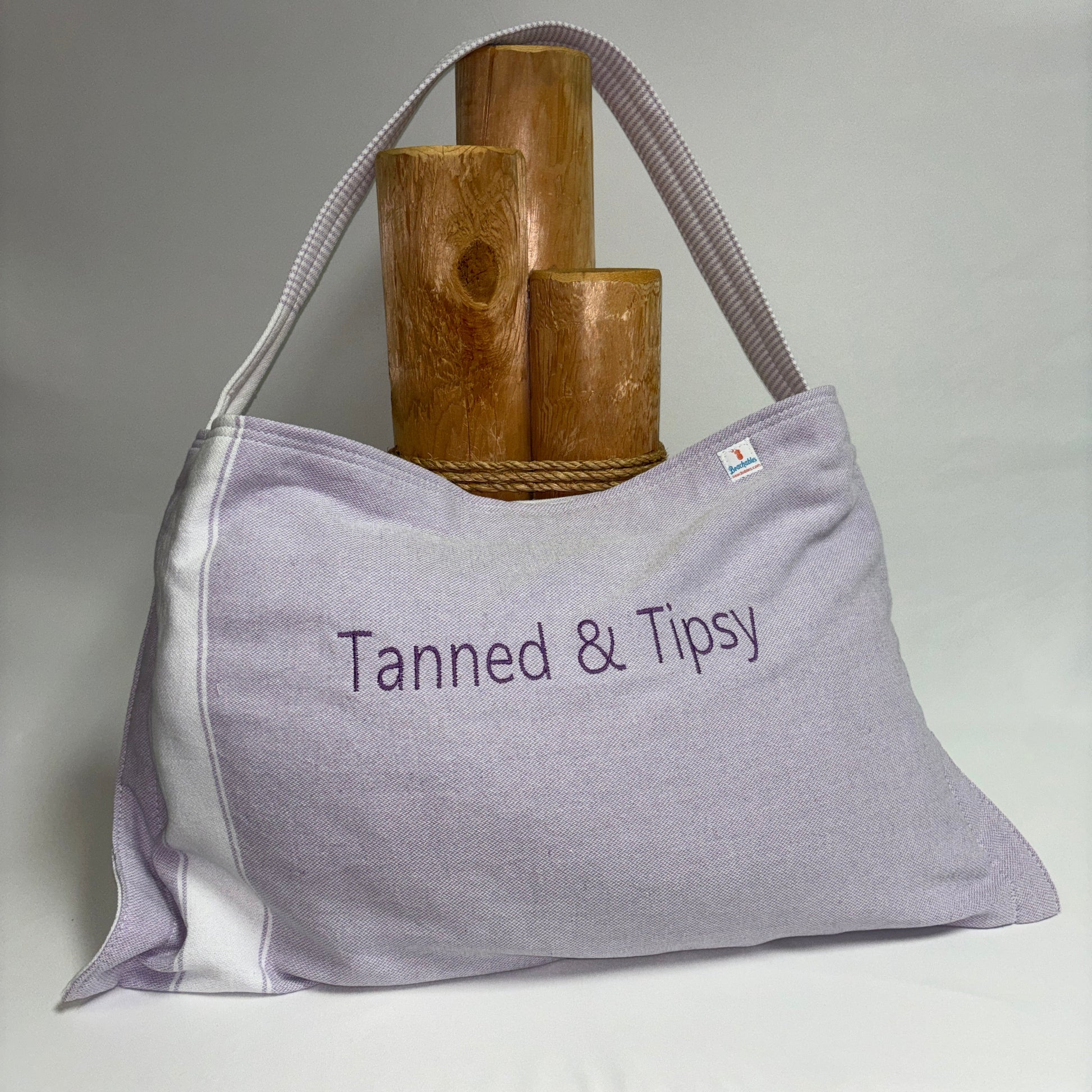 A light lavender tote bag with white side panels and a matching shoulder strap, featuring the embroidered text 'Tanned & Tipsy' on the front. It is styled with three wooden cylinder props on a white draped surface against a neutral gray background.