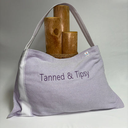 A light lavender tote bag with white side panels and a matching shoulder strap, featuring the embroidered text 'Tanned & Tipsy' on the front. It is styled with three wooden cylinder props on a white draped surface against a neutral gray background.