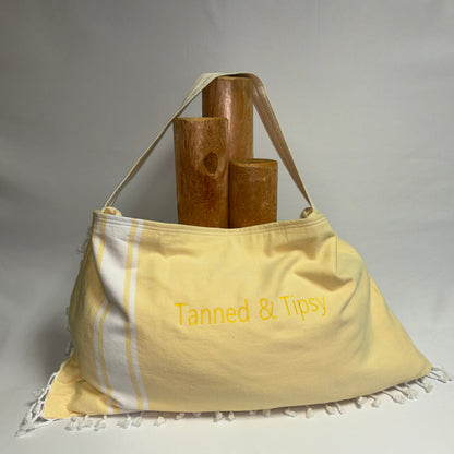 A pale yellow cotton towel bag with white stripes and white fringe, featuring the text 'Tanned & Tipsy' embroidered in yellow. The bag is styled with three wooden candle holders on top, set against a neutral gray fabric backdrop.