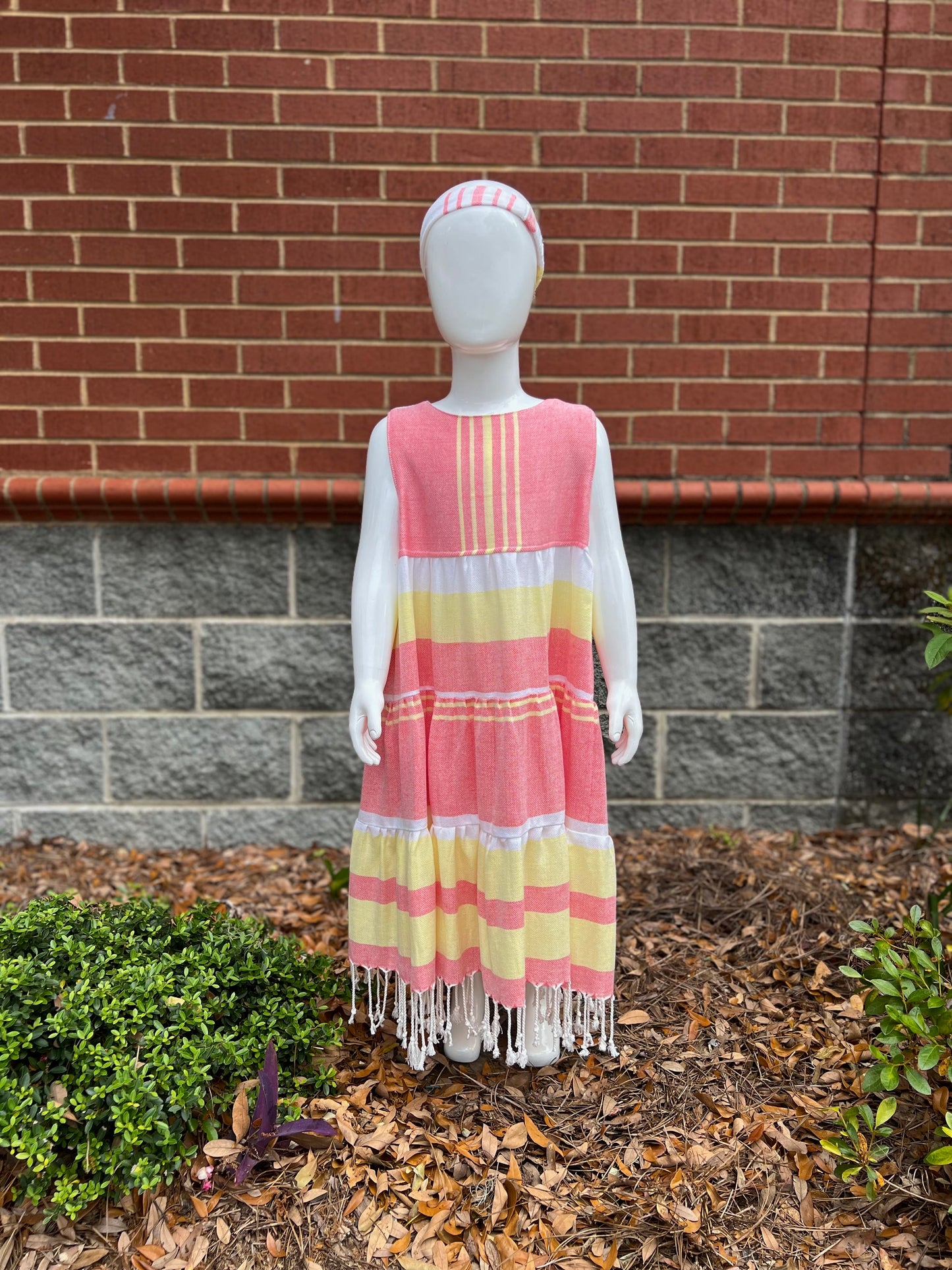 Mannequin wearing a three-tiered red, yellow and white striped sleeveless dress with tassels in front of a red brick wall