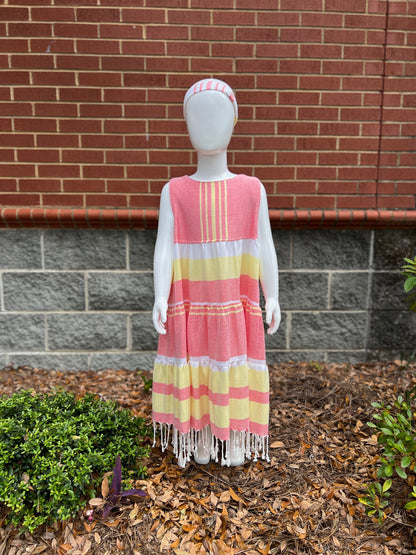 Mannequin wearing a three-tiered red, yellow and white striped sleeveless dress with tassels in front of a red brick wall
