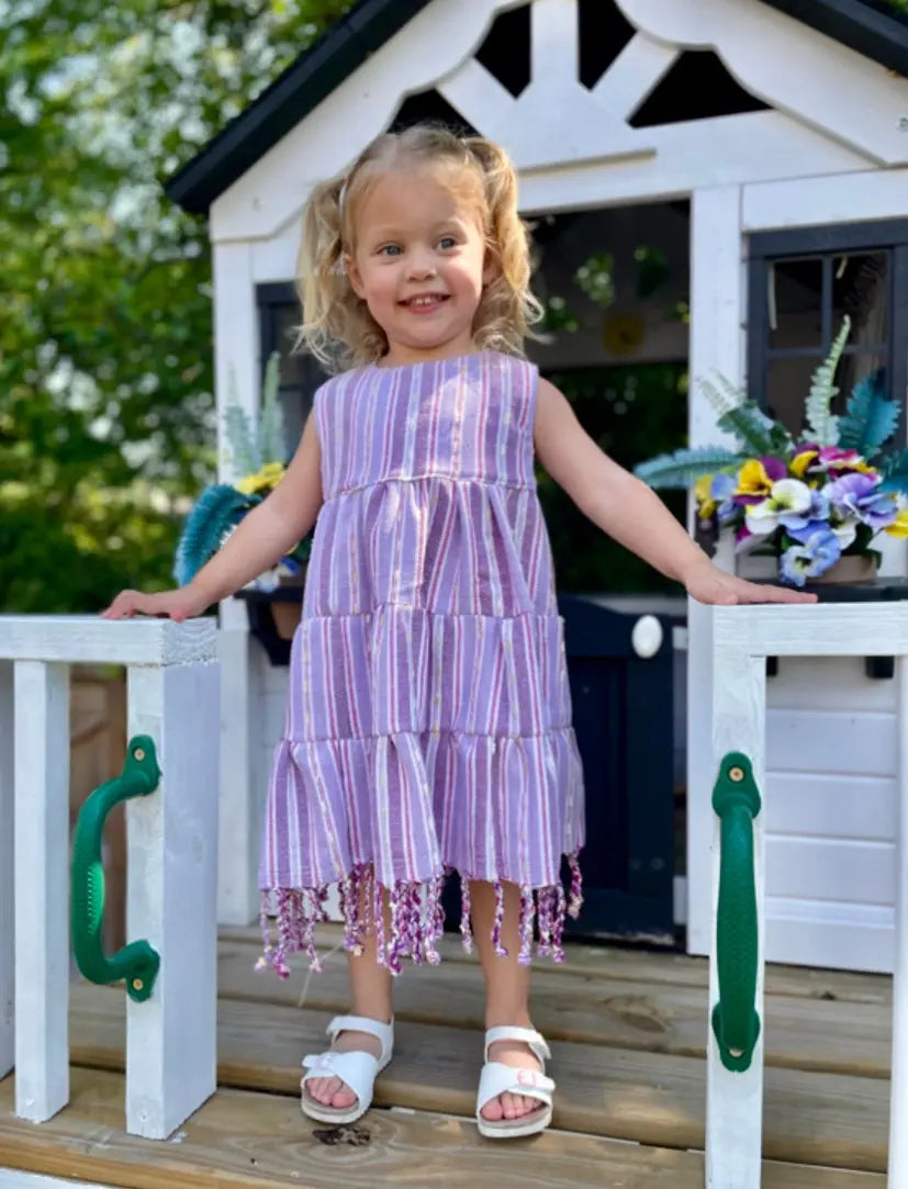 Young girl in a purple three tiered, striped dress with tassels standing in front of a white playhouse with green handles.