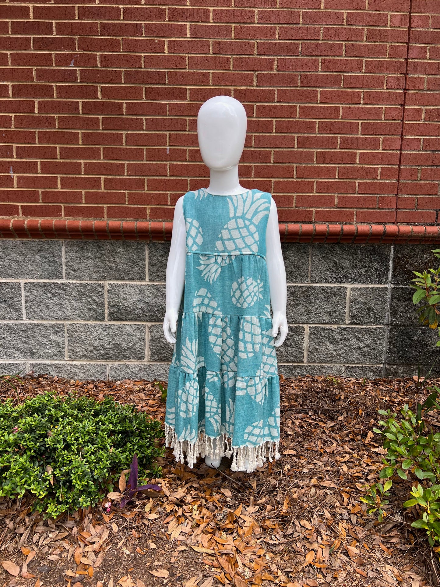 Mannequin wearing a three-tiered green pineapple print sleeveless dress with tassels in front of a red brick wall