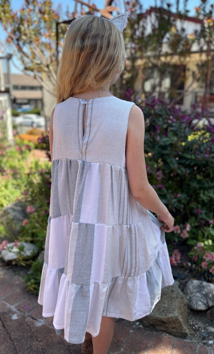 Back view of a young girl wearing a three-tiered grey, charcoal and white striped sleeveless dress with tassels outside