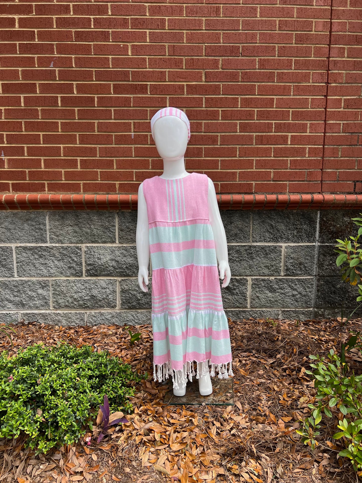 Mannequin wearing a three-tiered pink, green and white striped sleeveless dress with tassels in front of a red brick wall