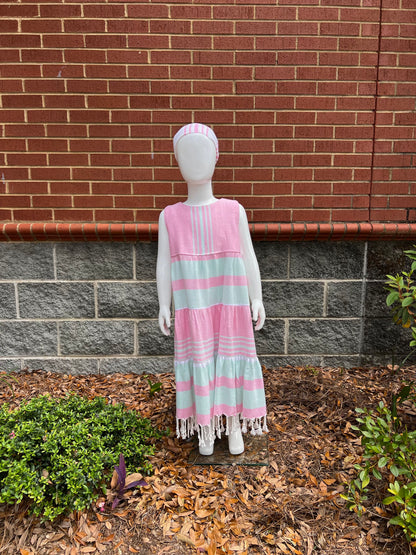 Mannequin wearing a three-tiered pink, green and white striped sleeveless dress with tassels in front of a red brick wall