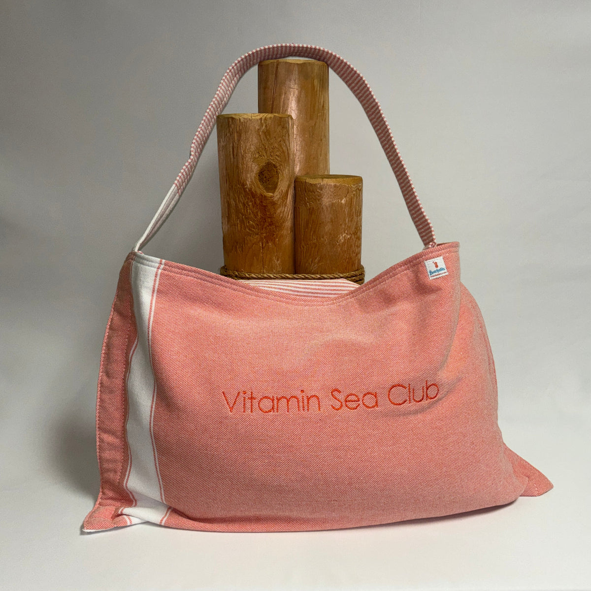 A pink canvas shoulder bag with white side panels and a pink-and-white striped handle, featuring the embroidered text 'Vitamin Sea Club' on the front. The bag is styled with three wooden cylindrical props on a draped white fabric surface against a neutral gray backdrop.