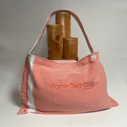 A pink canvas shoulder bag with white side panels and a pink-and-white striped handle, featuring the embroidered text 'Vitamin Sea Club' on the front. The bag is styled with three wooden cylindrical props on a draped white fabric surface against a neutral gray backdrop.