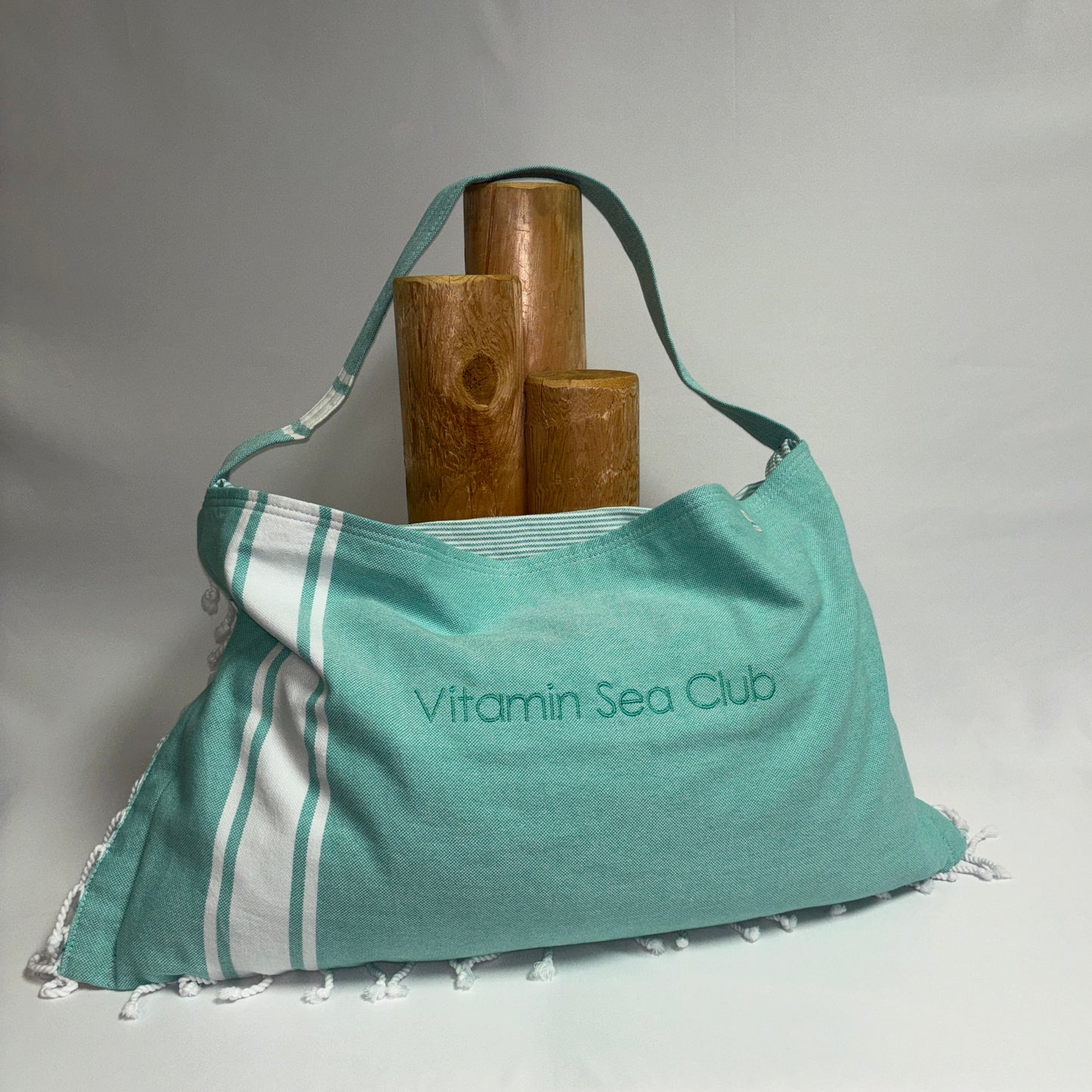 A teal and white striped beach towel shoulder bag with 'Vitamin Sea Club' printed on the front, styled with three wooden candle holders against a white fabric backdrop.