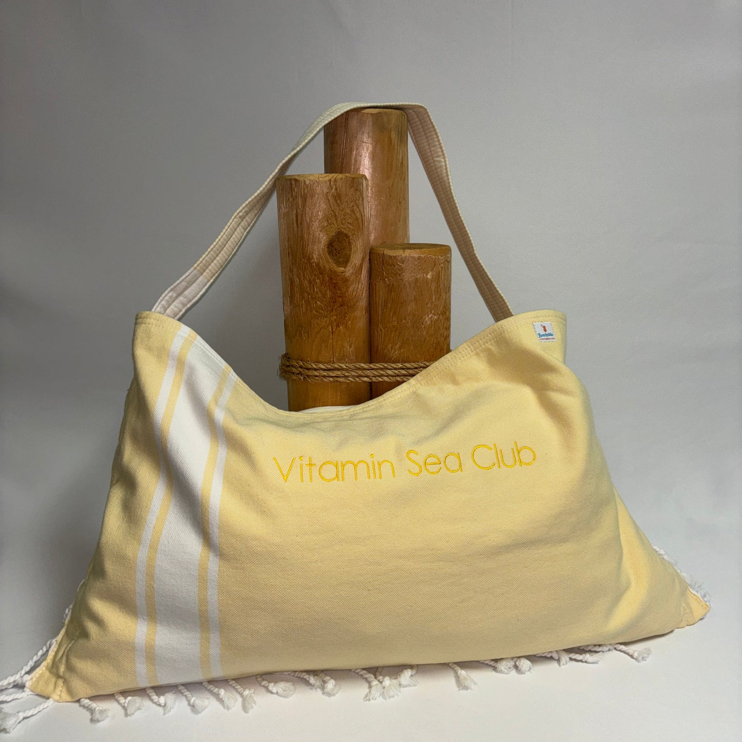 A pale yellow shoulder bag with white stripes and fringed edges, featuring the text 'Vitamin Sea Club' printed in yellow on the front. The bag is styled with a rope and three wooden poles against a neutral gray background.