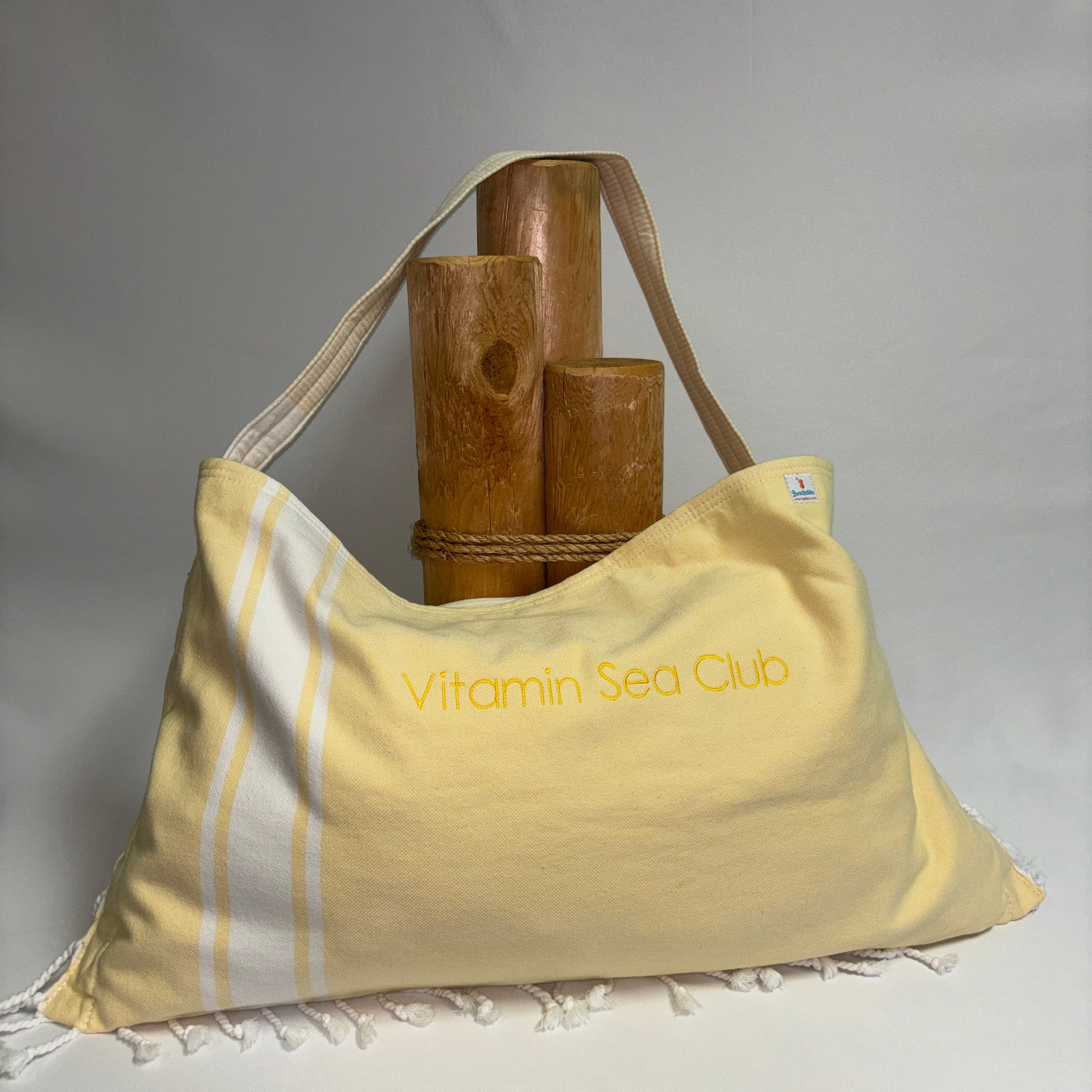 A pale yellow shoulder bag with white stripes and fringed edges, featuring the text 'Vitamin Sea Club' printed in yellow on the front. The bag is styled with a rope and three wooden poles against a neutral gray background.