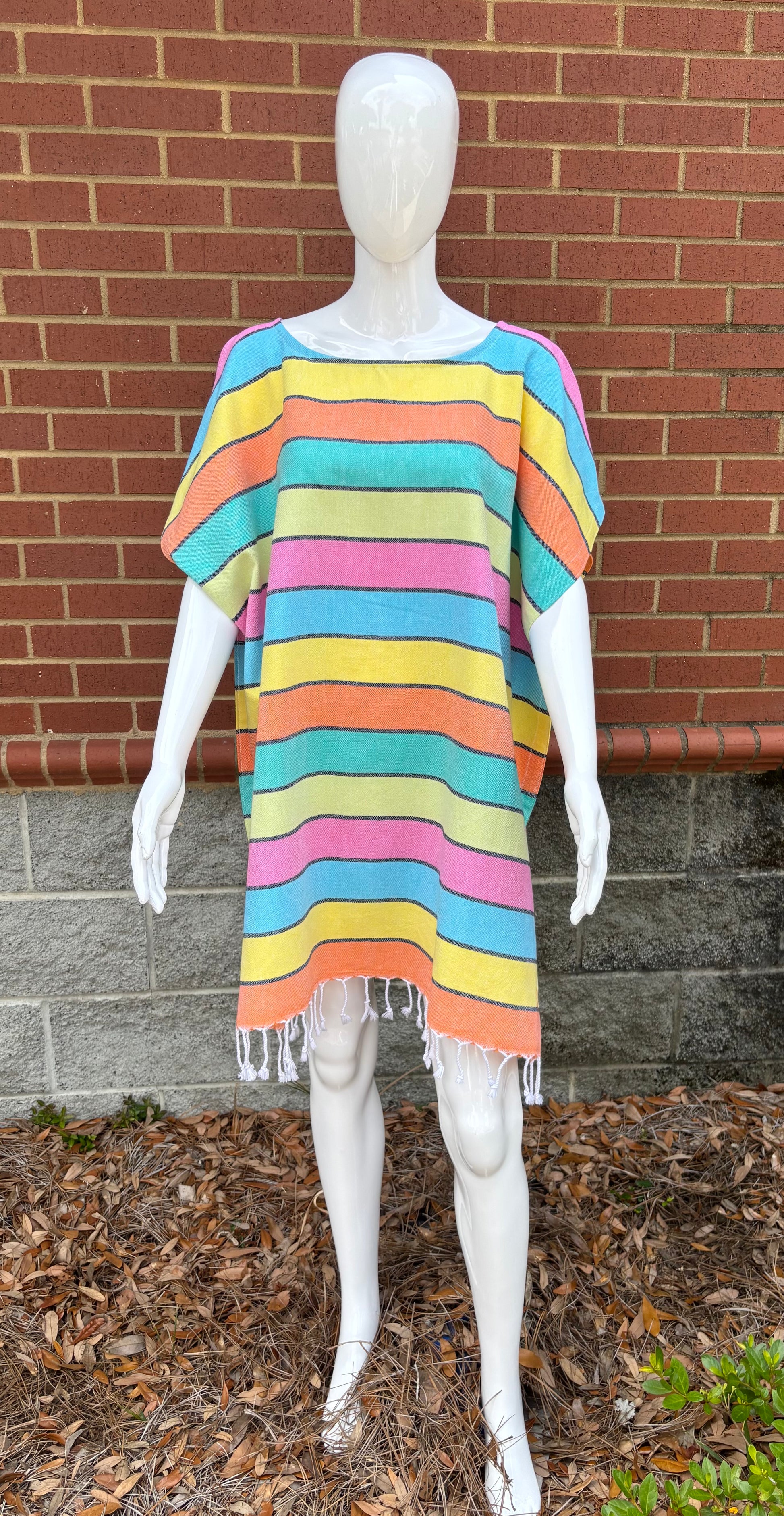 A mannequin displaying a women's poncho in orange, turquoise,  yellow, pink and teal stripes, with a brick wall and plants in the background.